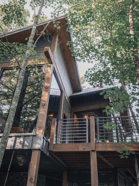 An exterior view of a modern cabin built on tall wooden stilts among green trees, showing a large glass window, a wooden deck with metal railings, and natural surroundings. Architecture by James Knight Photography by Kalyn Kight. Reynard Architectural Designs
