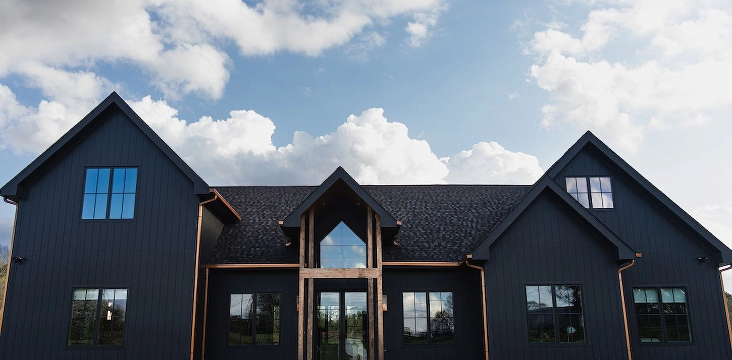A symmetrical modern farmhouse style home with striking matte black vertical siding and a black shingle roof under a blue sky with puffy white clouds. The central entryway features a high peaked roof supported by natural wood pillars. Powder Springs, GA