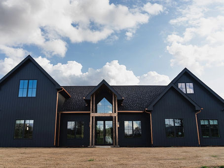 Dark gray modern farmhouse with gabled roof and wood entry, set in a grassy yard with spring trees.