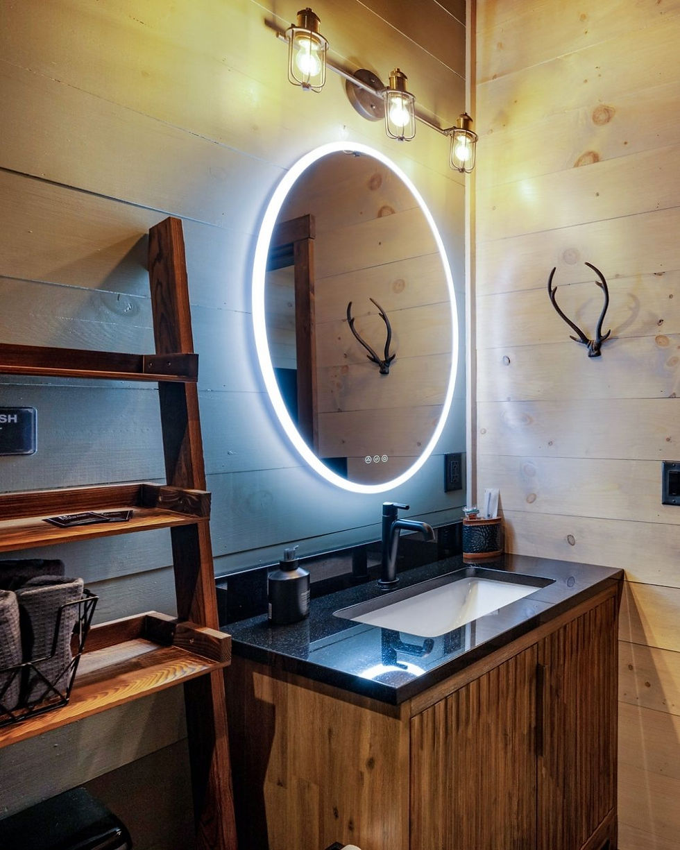 Rustic bathroom with wood paneling, illuminated round mirror, black countertop, and antler decor. Warm lighting creates a cozy mood. Reynard Architectural Designs