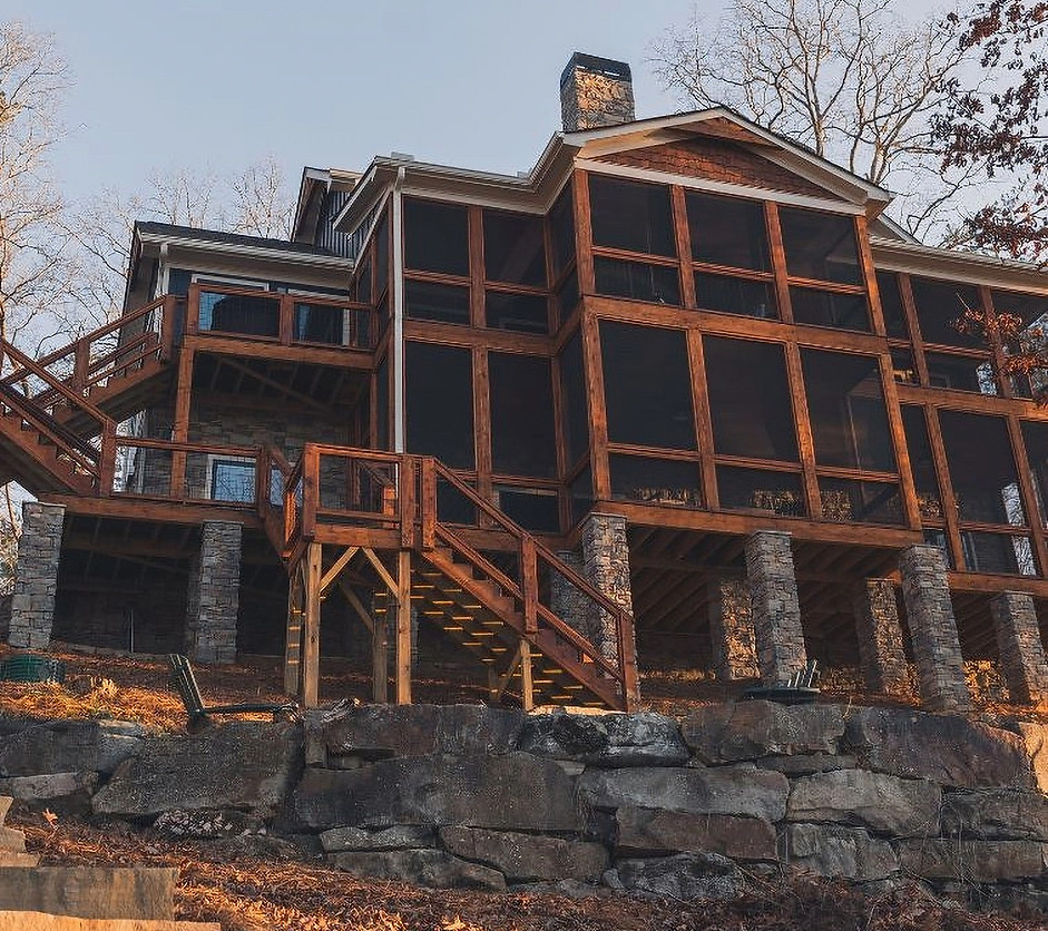 Alt Text: Low-angle rear exterior view of a multi-story modern rustic lakehouse built on a steep slope. The home features expansive, stacked screened porches with rich timber framing, supported by tall natural stone columns. A wooden staircase descends over a rugged retaining wall built from large natural boulders, bathed in warm, late-afternoon sunlight. Reynard Architectural Designs in Blairsville, GA on Lake Nottely. Custom Home.