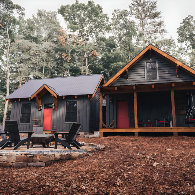 A stone patio fire pit area with dark Adirondack chairs set in a yard of reddish-brown mulch. In the background are two connected, small modern dark-paneled cabins with bright red front doors.Spring Camp. Ellijay, GA Reynard Architectural Designs