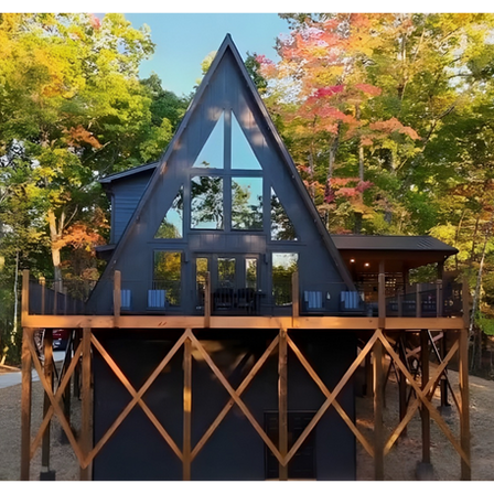 Modern black A-frame cabin featuring a large triangular window wall, an elevated deck, and unique X-braced wooden supports, set against a backdrop of autumn trees. Reynard Custom Homes