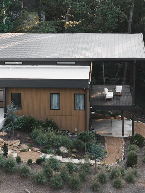 Aerial view of the home's right wing showing warm wood exterior siding, a large covered balcony with two white lounge chairs, and an under-deck patio area surrounded by landscaped terrain. Architecture by James Knight Photo by Kalyn Kight Reynard Architectural Designs