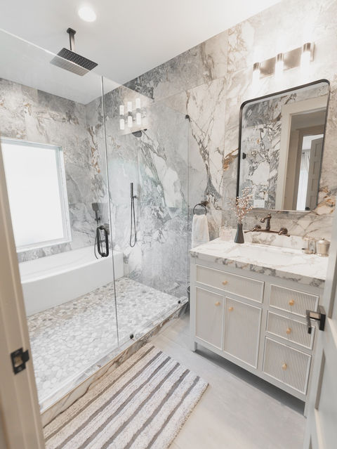 Modern bathroom with a glass-enclosed walk-in shower featuring large gray veined tiles and a pebble floor, next to a light gray fluted vanity with a marble countertop and black fixtures. Architecture by James Knight Photo by Kalyn Kight Reynard Architectural Designs
