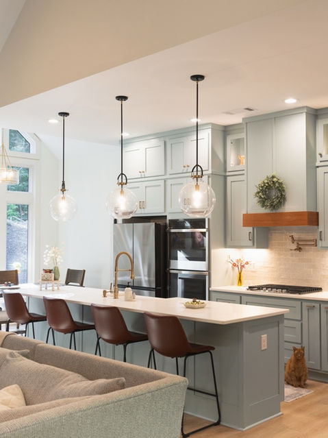 Open-concept living space showing the back of a grey sofa, a kitchen with a white island and light blue cabinets, a dining area with large windows, and an orange cat sitting on the kitchen floor. Custom home designed by Reynard Architectural Designs