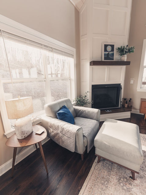 Interior view of a cabin living room showing a tall white paneled fireplace reaching toward a vaulted wood-plank ceiling, next to a large window and gray armchair. Mountain home architecture by Reynard Architectural Designs