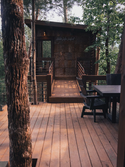 A wooden deck area with a dining table leading to a short wooden bridge that connects to the entrance of a cabin covered in natural bark siding, built seamlessly among pine trees. Architecture by James Knight Photography by Kalyn Kight. Reynard Architectural Designs