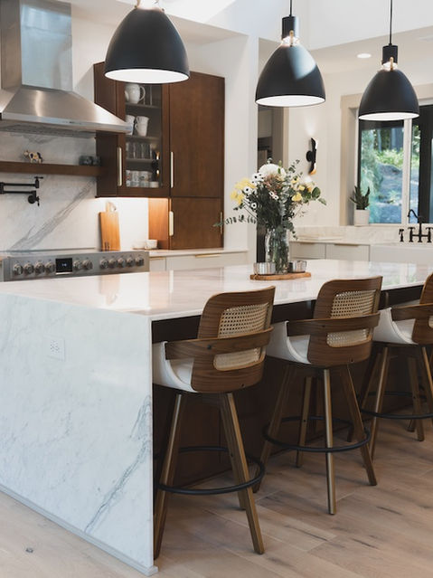 Modern kitchen featuring a white marble waterfall island with three wood and cane barstools, dark wood upper cabinets, white lower cabinets, and three large black pendant lights. Architecture by James Knight Photo by Kalyn Kight Reynard Architectural Designs