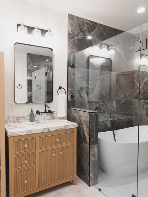 Modern bathroom showcasing a light wood fluted vanity with a marble top, matte black fixtures, and a glass-enclosed wet room containing a freestanding white tub and dark gray veined wall tiles. Architecture by James Knight Photo by Kalyn Kight Reynard Architectural Designs