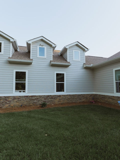 Side exterior view of a home showcasing three roof dormer windows, light grey horizontal siding, and a decorative stone base. Architecture by James Knight. Photo by Kalyn Kight