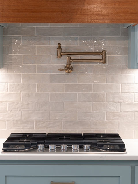 Close-up of a kitchen featuring a black gas cooktop, a light glossy subway tile backsplash, a bronze pot filler, and light blue cabinetry. Custom home designed by Reynard Architectural Designs