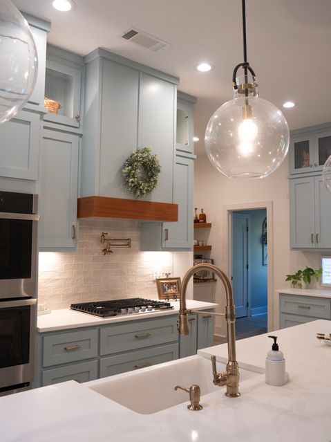 White kitchen island countertop with an undermount sink and brass faucet, looking past light blue cabinets and a gas range toward an open hallway. Custom home designed by Reynard Architectural Designs
