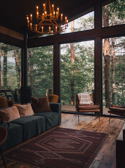 A cabin living room featuring a dark green plush sofa, a brown patterned area rug, leather accent chairs, a black freestanding wood stove, and expansive windows looking out into a green forest. Architecture by James Knight Photography by Kalyn Kight. Reynard Architectural Designs