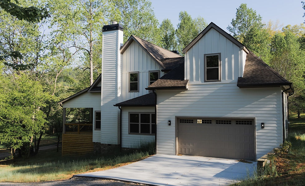 Exterior of a custom modern mountain home in Hiawassee, GA featuring white siding, a prominent chimney, a semi-attached garage, and a covered wood porch surrounded by a lush forest. Reynard Architectural Designs 