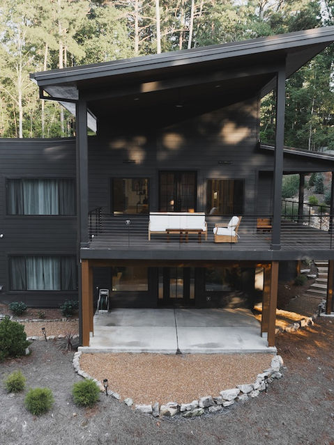 Aerial view of a modern dark gray home's rear exterior featuring a sloped roof, a large balcony with white outdoor furniture, wood support beams, and a terraced stone walkway. Architecture by James Knight Photo by Kalyn Kight Reynard Architectural Designs