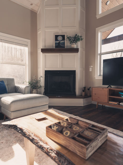 Cozy cabin living room interior featuring a white paneled corner fireplace, a light gray armchair with an ottoman, and a wooden coffee table. Mountain home architecture by Reynard Architectural Designs