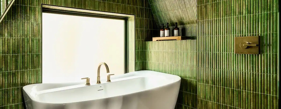 Bathroom Retreat: A luxurious spa-like bathroom featuring a freestanding tub surrounded by green tiled walls and brass fixtures, set under the A-frame's sloped ceiling. Photographed by Moth Rust Media, with interior designs by Reynard Custom Homes.