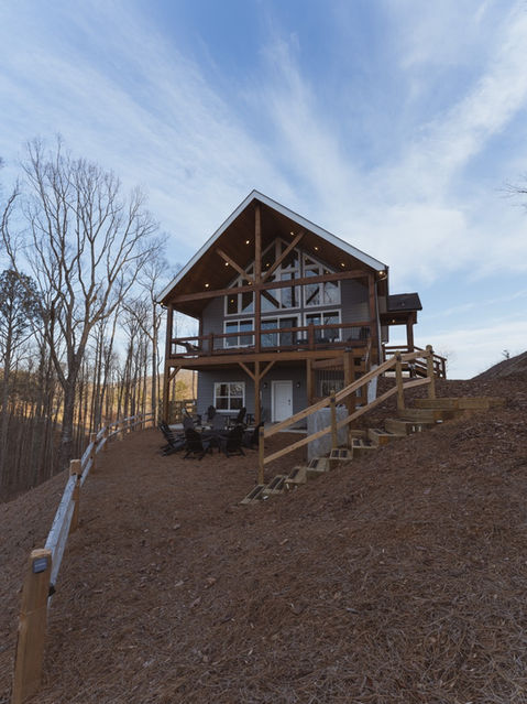 Angled exterior view of a two-story cabin featuring a lower patio with a fire pit, an upper deck, and wooden stairs climbing the hill beside the home. Mountain home architecture by Reynard Architectural Designs