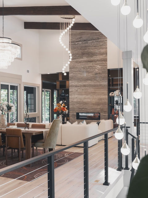View from an upper landing looking down into an open-concept living and dining area, featuring a tall stone fireplace, cascading glass chandeliers, dark ceiling beams, and a black wire railing in the foreground. Architecture by James Knight Photo by Kalyn Kight Reynard Architectural Designs