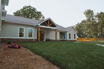 Low-angle exterior view of a single-story home featuring light grey siding and a stone veneer foundation. A gabled portico with natural wood columns and truss details frames the dark double-door entrance. In the foreground, bright pink flowers bloom in a pine straw bed bordering a lush green lawn, while a yellow wooden fence is visible in the background to the right. Reynard Custom Homes Cumming GA Architect James Knight.jpeg
