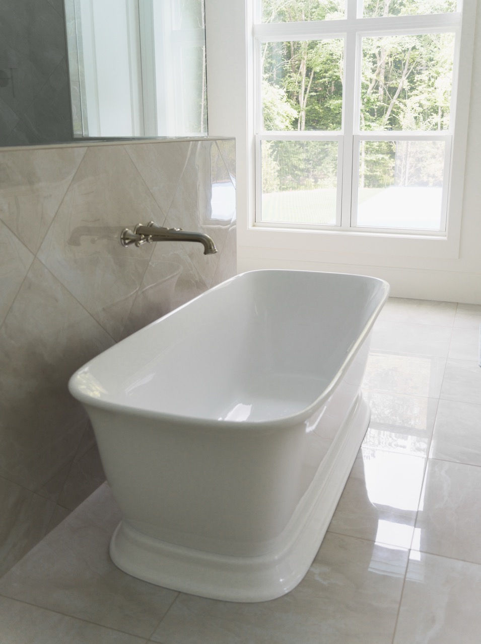 Bright modern bathroom with a white freestanding bathtub, wall faucet, beige tile, and a large window looking onto trees. Photo by Kalyn Kight. Architecture by James Knight (Reynard Architectural Designs)