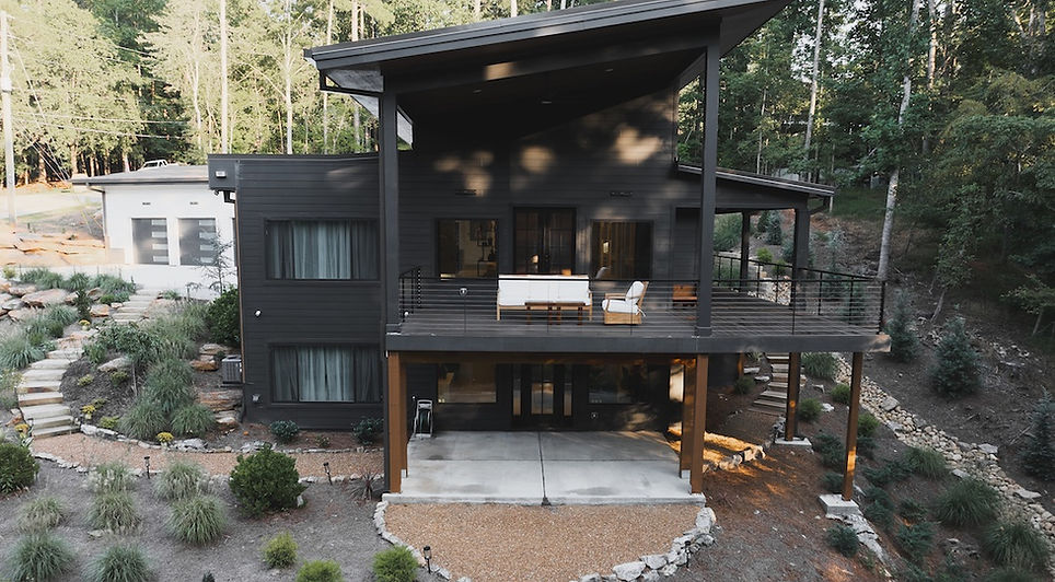 An aerial view of a modern, two-story dark charcoal house with a dramatic slanted roof, featuring a large upper deck with outdoor furniture and a beautifully landscaped, sloped yard surrounded by trees.