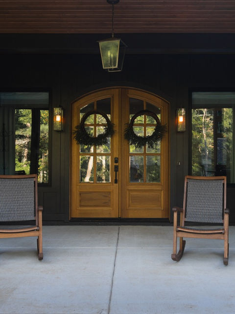 Covered front porch with dark grey siding, symmetrical wooden rocking chairs, and arched wooden double front doors decorated with green wreaths. Reynard Architectural Designs