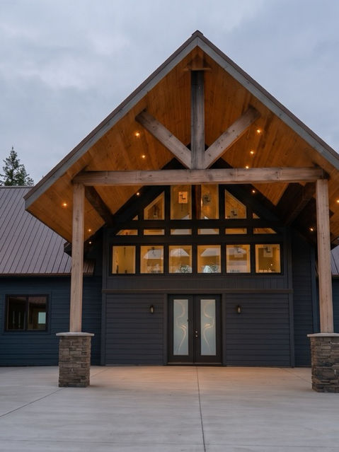 Custom mountain estate front entrance featuring a large gabled timber-frame porte-cochère, stone-based wooden posts, dark siding, double front doors, and a spacious concrete driveway. Talking Rock, GA Reynard Architectural Designs
