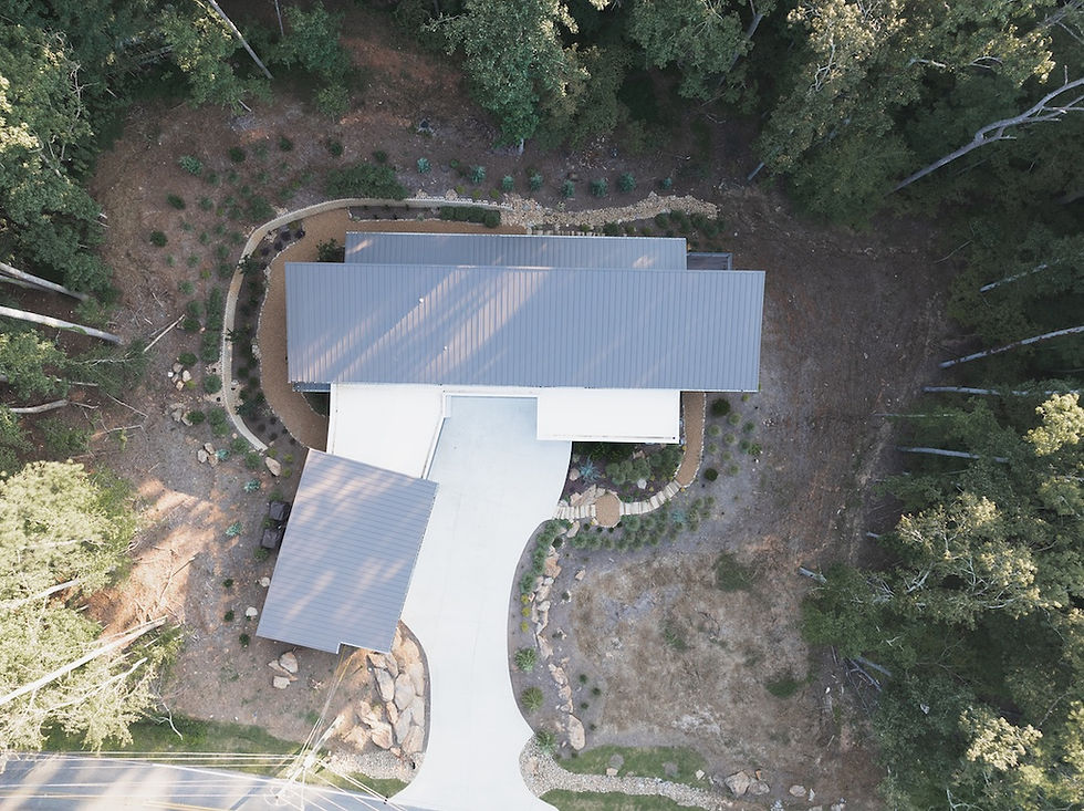 Aerial view of a modern house with a gray roof, surrounded by trees and landscaped garden. Curved driveway leads to the entrance. Reynard Custom Homes architecture