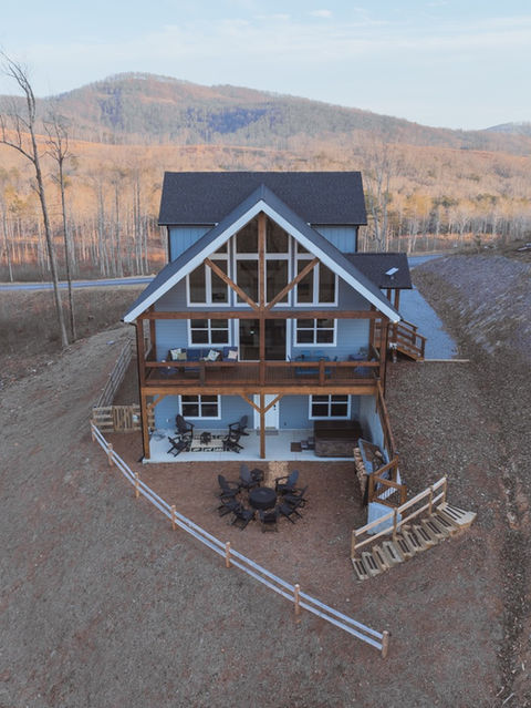 Aerial view of a modern cabin's rear elevation featuring large windows, multiple outdoor living spaces, and a split-rail fence on a sloped dirt hillside. Mountain home architecture by Reynard Architectural Designs