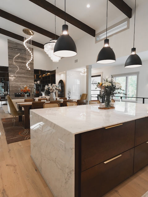 Wide view of an open-concept living space highlighting a large marble kitchen island with dark wood drawers, leading into a dining area and a living room with a tall stone fireplace. Architecture by James Knight Photo by Kalyn Kight Reynard Architectural Designs
