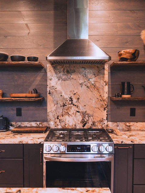 A kitchen interior featuring a stainless steel gas range, dramatic stone backsplash, floating wooden shelves decorated with rustic bowls, and dark gray cabinets against wood-paneled walls. Architecture by James Knight Photography by Kalyn Kight. Reynard Architectural Designs