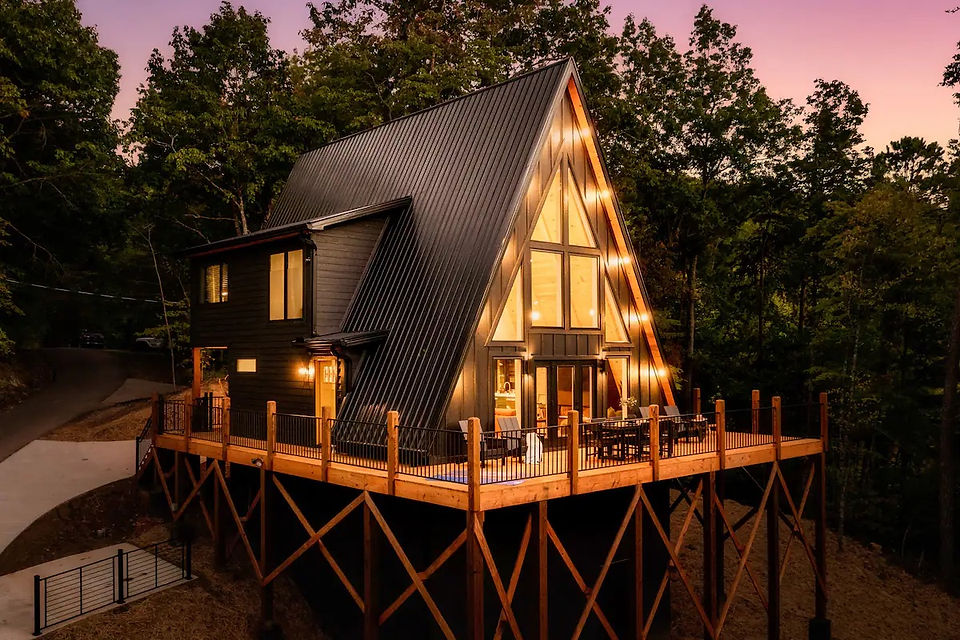 An enchanting A-frame home glowing at dusk, set against the backdrop of the lush Great Smoky Mountains. The elevated wooden deck wraps around the home, offering expansive outdoor living spaces. Photo by Moth Rust Media. Architectural and interior design by Reynard Custom Homes.