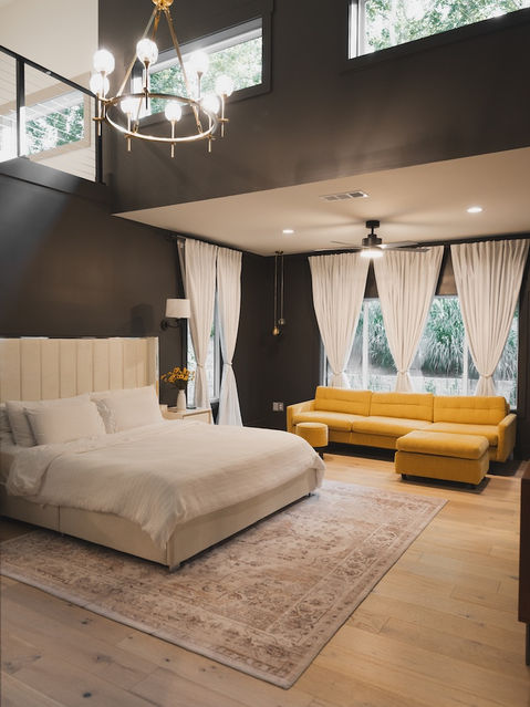 Large primary bedroom featuring dark walls, high ceilings with a brass chandelier, a cream-colored upholstered bed, a bright yellow modern sectional sofa, and light hardwood floors. Architecture by James Knight Photo by Kalyn Kight Reynard Architectural Designs