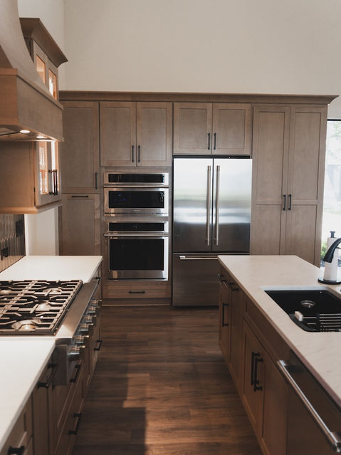 A kitchen wall featuring warm wood, flat-panel cabinets seamlessly integrating a stainless steel refrigerator and stacked double wall ovens. A kitchen island with a white countertop and black sink sits in the foreground. Architecture by James Knight. Photo by Kalyn Kight (Reynard Architectural Designs)