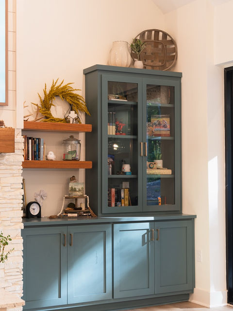 Custom teal built-in cabinets with glass upper doors and two wooden floating shelves decorated with a yellow wreath and books beside a stone fireplace. Custom home designed by Reynard Architectural Designs