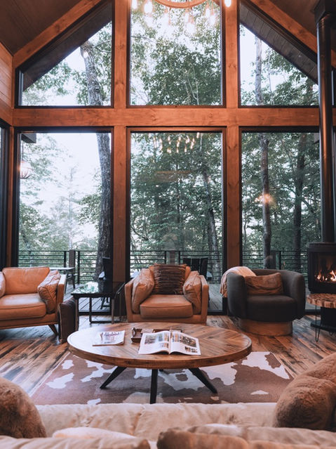 A straight-on view of a living room with massive A-frame windows revealing a dense forest, featuring a round wooden coffee table, a dark sofa, leather chairs, and a corner wood stove. Architecture by James Knight Photography by Kalyn Kight. Reynard Architectural Designs