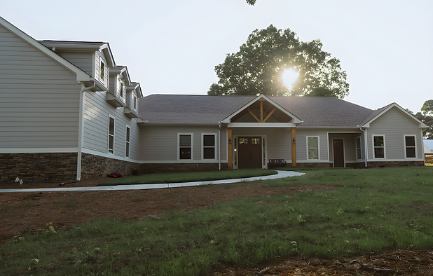 A wide front view of a sprawling single-story beige house with horizontal siding and a stone-veneer foundation. A white concrete walkway curves across a grassy yard toward the central front porch under a bright sky with the sun peeking behind a large tree. Cumming, GA