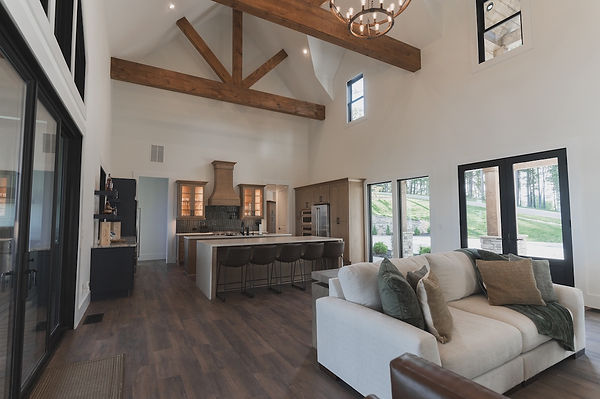 Spacious custom master bedroom with vaulted ceilings and thick exposed wood beams. The room features dark wood flooring, a bed positioned under a black-framed window, and unique illuminated built-in shelving and cabinetry integrated into the sloped roofline. Reynard Custom Homes.