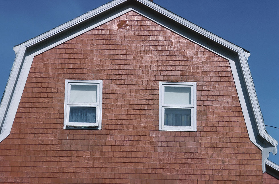 Close-up of a house a gambrel roof with reddish wood shingles, two white-framed windows, and a clear blue sky above. Reynard Architectural Designs