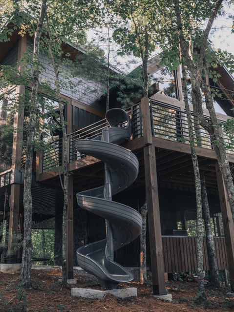 An upward-facing exterior shot of a modern treehouse-style cabin elevated on wooden pilings among tall green trees. A large, dark gray spiral tube slide connects the upper wooden deck to the pine-needle-covered forest floor below. Architecture by James Knight Photography by Kalyn Kight. Reynard Architectural Designs