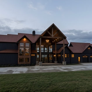 Front exterior view of a custom dark board-and-batten modern lodge in Talking Rock, GA, designed by Reynard Architectural Designs, featuring a timber frame entry portico with stone pillars, a rust-colored metal roof, and lighted floor-to-ceiling windows.