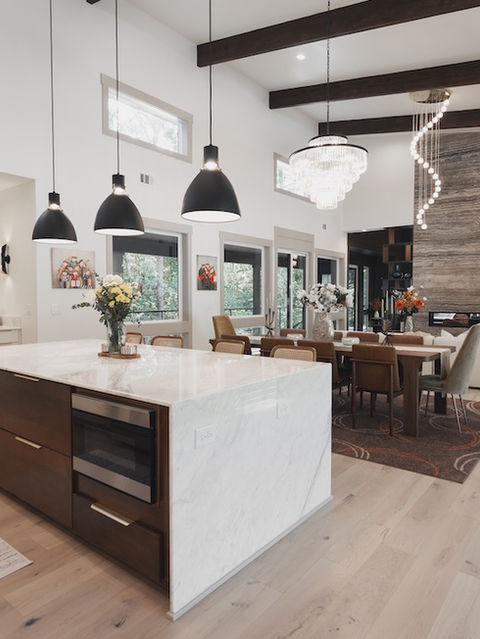 Wide shot of a modern open-concept kitchen and living room featuring a white marble waterfall island, dark pendant lights, light hardwood floors, and a tall stone fireplace. Architecture by James Knight Photo by Kalyn Kight Reynard Architectural Designs