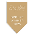 A muted bronze, banner-shaped award badge featuring white text. The top reads "Design Skill Awards" with "Design Skill" written in a cursive script and "Awards" in small capital letters. The bottom section reads "BRONZE WINNER 2025" in bold, sans-serif capital letters. Reynard Custom Homes Architecture