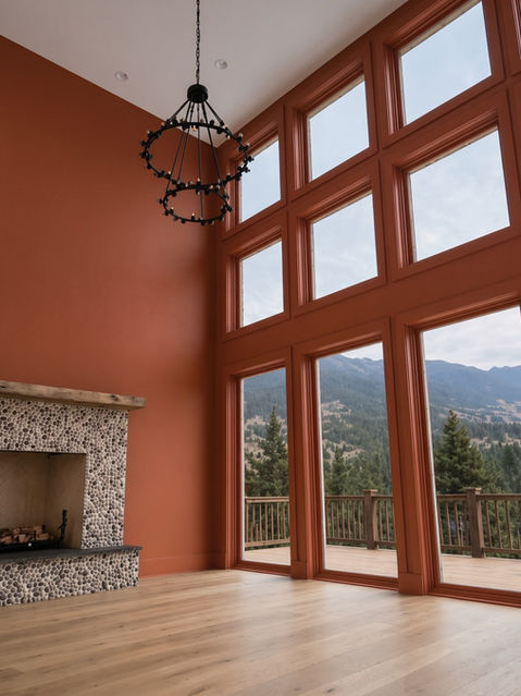 High-ceiling living room with terracotta painted walls, river pebble fireplace, light oak wood floors, black metal chandelier, and large window wall displaying mountain views. Talking Rock, GA Reynard Architectural Designs