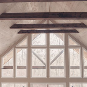 Upward interior view of a cabin's vaulted light-wood ceiling accented with dark timber beams and a large multi-paned A-frame window looking out to bare trees.