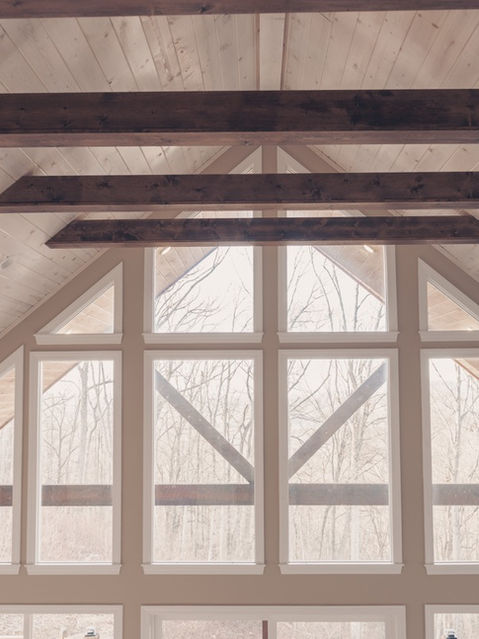 Upward interior view of a cabin's vaulted light-wood ceiling accented with dark timber beams and a large multi-paned A-frame window looking out to bare trees. Mountain home architecture by Reynard Architectural Designs