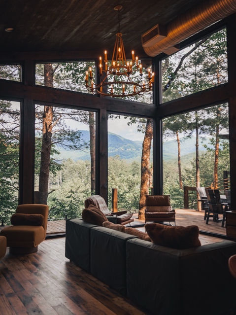 A spacious living room with large A-frame windows offering a view of lush green mountains, featuring a plush dark sofa, leather chairs, and a large multi-tier candle-style chandelier.
Architecture by James Knight Photography by Kalyn Kight. Reynard Architectural Designs