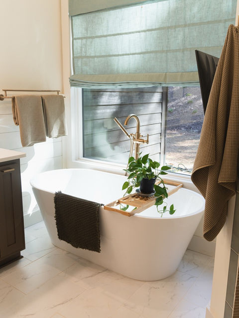 White freestanding bathtub with a wooden bath caddy and potted plant, positioned in front of a large window with a light blue roman blind, featuring brass hardware and light marble-look floor tile.